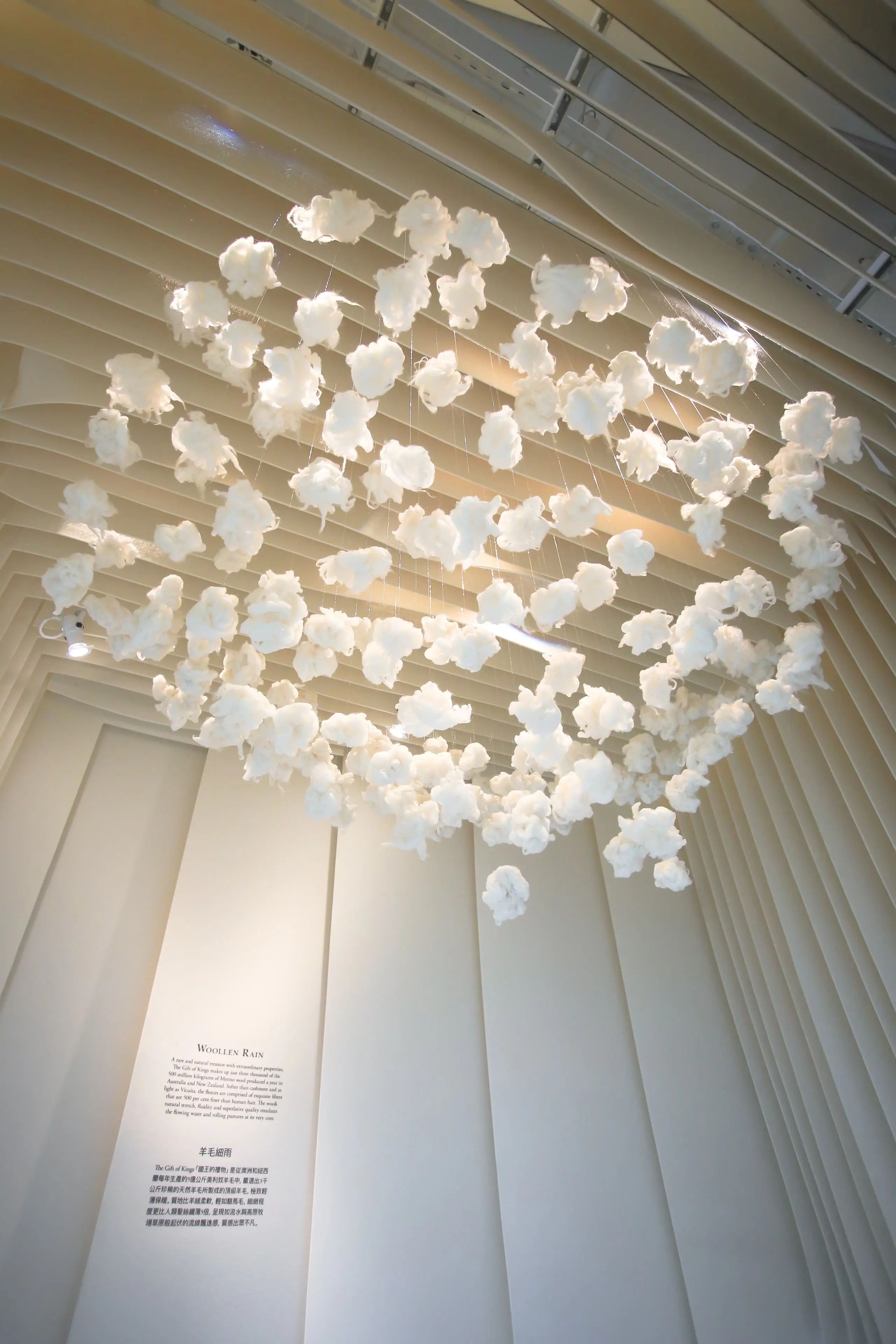 Clusters of soft, cloud-like wool forms suspended from the ceiling above an exhibition space, creating a floating installation effect.